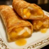Crispy golden-brown peach cobbler egg rolls dusted with cinnamon sugar and drizzled with vanilla glaze on a white serving plate