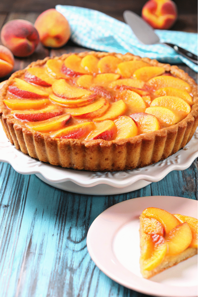 Golden brown peach tart recipe with overlapping peach slices arranged in concentric circles, topped with crumbly butter mixture, served on white plate with dollop of whipped cream