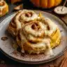 Golden brown pumpkin butter cinnamon rolls in a baking dish, covered with creamy maple cream cheese glaze, photographed from above on a rustic wooden surface with fall leaves and a cup of coffee in the background.