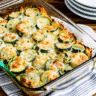 Golden Cheesy Zucchini Bake in white baking dish with melted cheese on top and fresh basil leaves scattered around