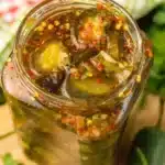 Colorful jar of sweet heat pickles with jalapeño slices and red pepper flakes visible through the glass, sitting on a rustic wooden cutting board with fresh herbs scattered around