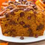 Golden brown chocolate chip pumpkin bread sliced on a wooden cutting board, showing moist interior with chocolate chips throughout, surrounded by autumn pumpkins and spices