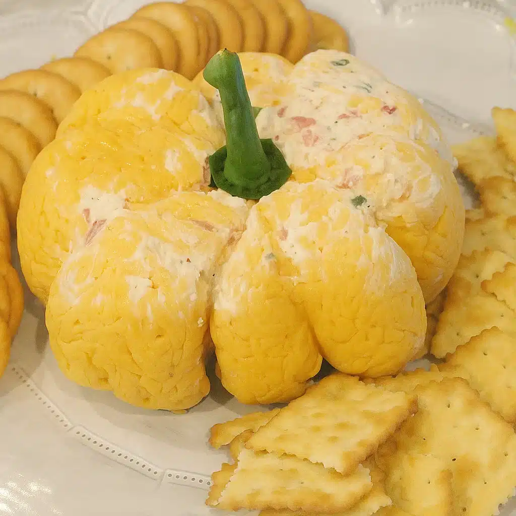 Pumpkin Cheese Ball combines shaped like a pumpkin with ridges and a red pepper stem, surrounded by crackers on a wooden board