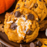 Golden brown pumpkin Smores cookies with melted marshmallow centers and chocolate chips, displayed on a rustic wooden board with fall leaves in the background
