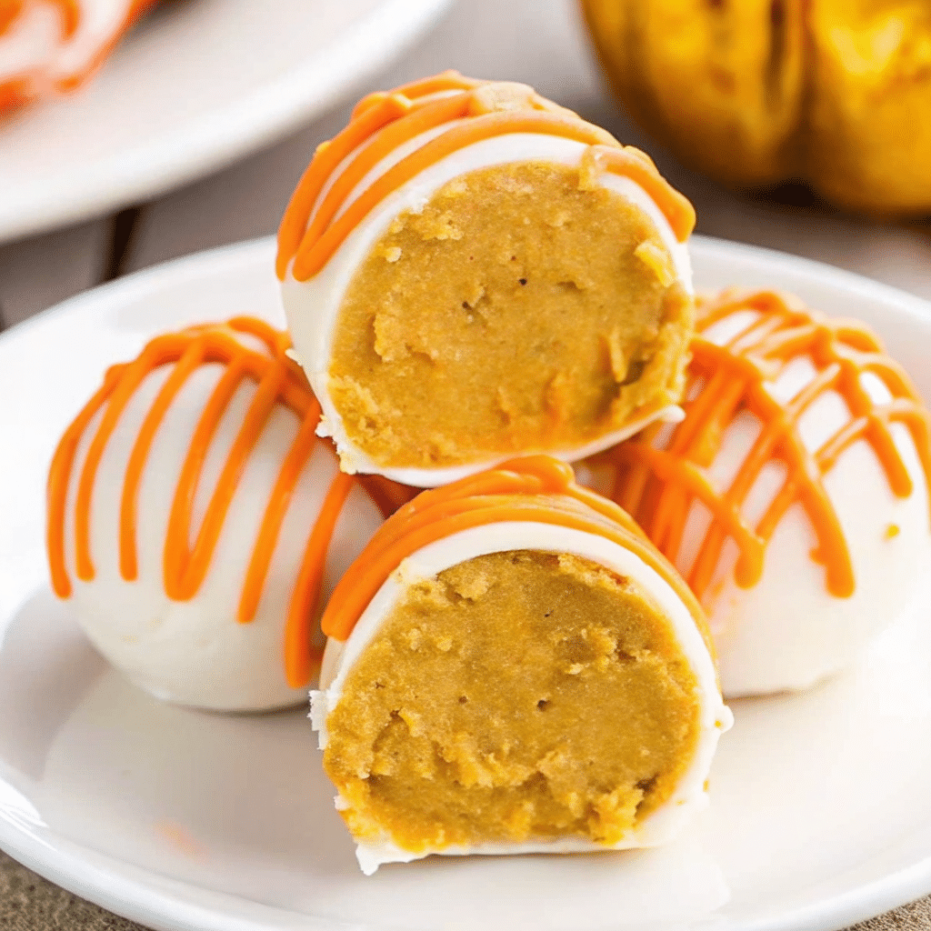 Bite-sized orange No Bake pumpkin cheesecake balls coated in smooth white chocolate with decorative orange drizzle, arranged on a white serving platter with fall leaves in the background