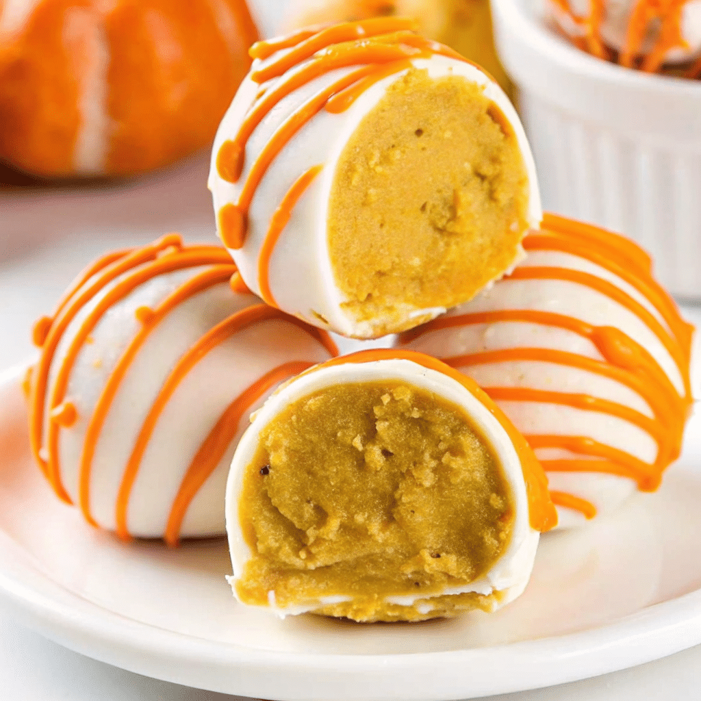 Bite-sized orange No Bake pumpkin cheesecake balls coated in smooth white chocolate with decorative orange drizzle, arranged on a white serving platter with fall leaves in the background