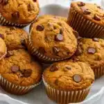 Golden brown pumpkin chocolate chip muffins topped with melted chocolate chips, arranged on a wire cooling rack with fall leaves in the background.