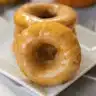 Golden brown pumpkin donuts with maple glaze dripping down the sides, arranged on a white plate with fall leaves in the background