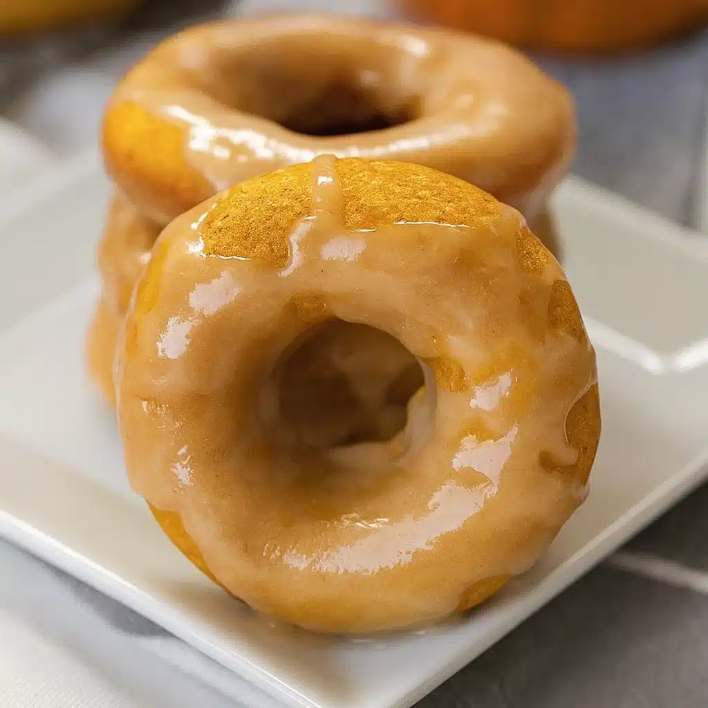 Golden brown pumpkin donuts with maple glaze dripping down the sides, arranged on a white plate with fall leaves in the background