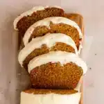 A slice of moist pumpkin bread with cream cheese frosting on a white plate, showing the beautiful orange color and fluffy texture, with fall spices scattered around
