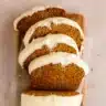 A slice of moist pumpkin bread with cream cheese frosting on a white plate, showing the beautiful orange color and fluffy texture, with fall spices scattered around
