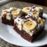 Square of banana pudding brownies on a white plate showing layers of chocolate brownie, vanilla pudding, sliced bananas, and whipped cream topping with vanilla wafer crumbs