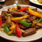 keto beef pepper steak stir-fry with tender sliced beef, red yellow and green bell peppers, garnished with sesame seeds and green onions in a glossy sauce on a white plate