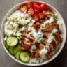 chicken shawarma bowl with sliced marinated chicken, jasmine rice, cherry tomatoes, cucumber, hummus, tzatziki sauce, and fresh parsley garnish