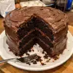 A slice of moist chocolate sour cream cake on a white plate with a fork, showing the tender crumb and rich dark color