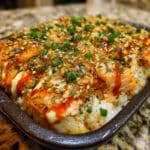 Spicy salmon sushi bake in a white baking dish topped with furikake, green onions, and drizzled with eel sauce and spicy mayo, served with roasted seaweed sheets