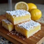 lemon bars dusted with powdered sugar, cut into squares and arranged on a white serving platter, showing the buttery shortbread crust and bright yellow lemon custard filling.