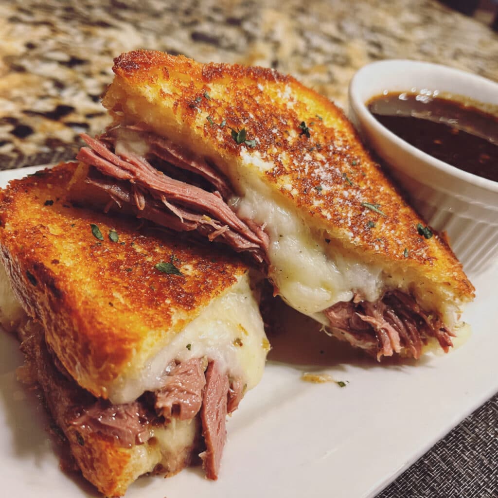 French Dip Grilled Cheese with roast beef and melted provolone cheese, cut in half and served on a plate next to a small bowl of warm brown au jus dipping sauce.