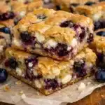 Square white chocolate blueberry blondies on a white plate showing golden edges, melted white chocolate throughout, and fresh blueberries baked into chewy bars