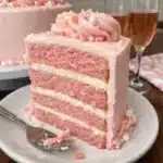 A three-layer pink champagne cake with smooth buttercream frosting, decorated with edible sparkles and pink sanding sugar on a white cake stand